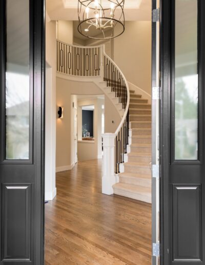 View from an open doorway into a foyer with a curved staircase, hardwood floors, and a modern chandelier.