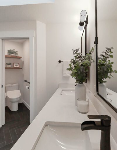 A modern bathroom featuring a white sink with a black faucet, large mirror, and an open door revealing a toilet and shelving unit. green potted plants add a touch of nature.