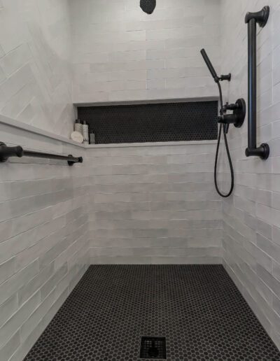 Modern accessible shower with white subway tiles, black hexagonal floor tiles, wall-mounted grab bars, handheld showerhead, and overhead rain shower.