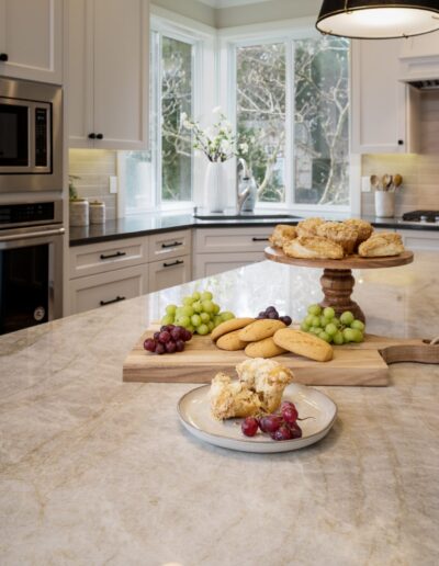 A modern kitchen with a marble countertop displaying grapes, cookies on a wooden stand, and a plate with a sliced pastry.