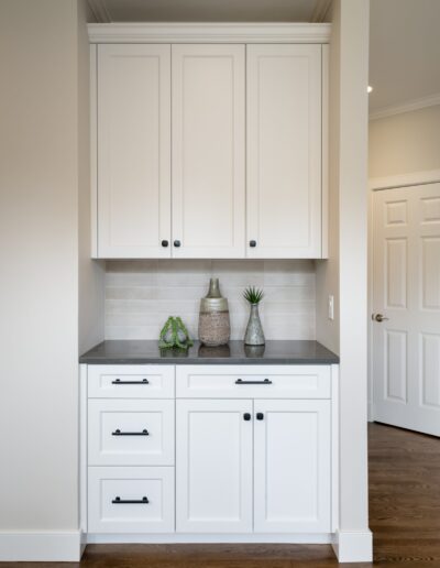 A modern kitchenette with white cabinets, black handles, a gray countertop, and decorative vases on the counter.