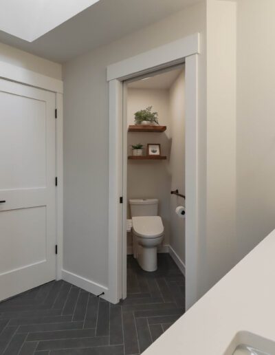 Modern bathroom with an open door revealing a toilet, white cabinetry, gray tiled floor, and a potted plant.