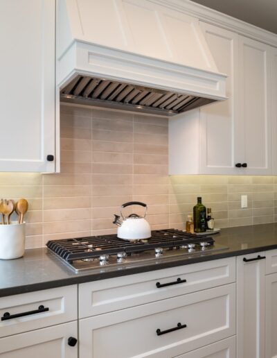 A modern kitchen with white cabinetry, a stainless steel stove, and a decorative range hood, complemented by a beige tile backsplash.