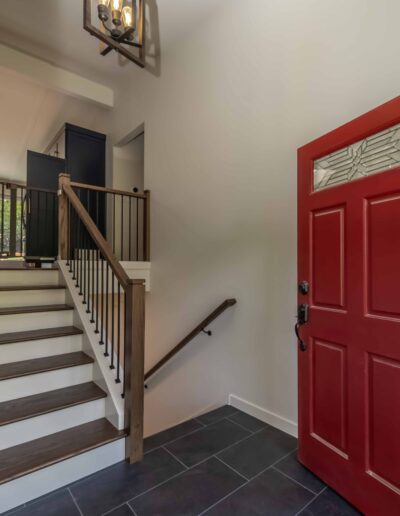 A brightly lit entryway with a red front door and a staircase leading to the upper floor, featuring a dark metal railing and a geometric pendant light fixture.