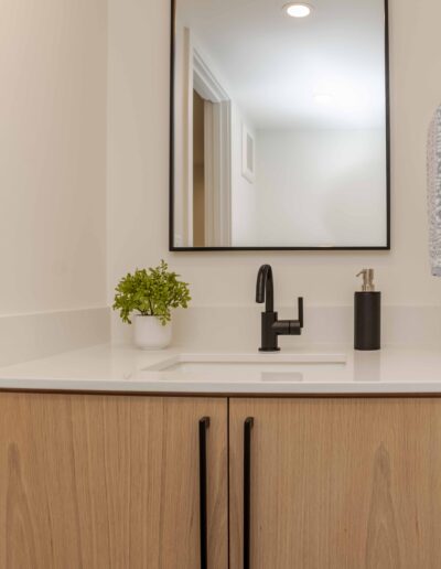 Modern bathroom vanity with a wooden cabinet, white countertop, black faucet, and rectangular mirror. A small potted plant and lotion dispenser are placed on the counter.