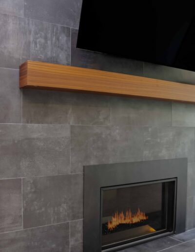 A modern fireplace with a sleek black frame is built into a dark gray tiled wall beneath a wooden mantel and a mounted flat-screen TV.