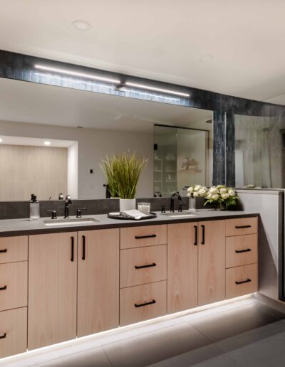 A modern bathroom featuring a large mirror, double sinks with a wooden vanity, and a glass-enclosed shower. The vanity has multiple drawers and there are shelves with towels and baskets.