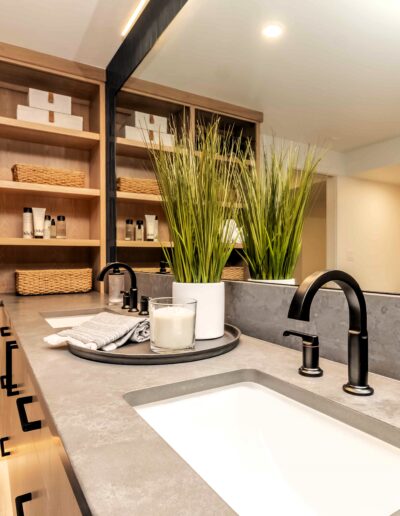 Modern bathroom with open shelving, rolled towels, wicker baskets, a large mirror, black fixtures, and a potted plant on a vanity with a grey countertop.