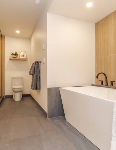 Modern bathroom with a freestanding bathtub, glass-enclosed shower, toilet, and wall-mounted shelf. Neutral tones with grey floor tiles and light wood accents. Bath towel hanging near the tub.