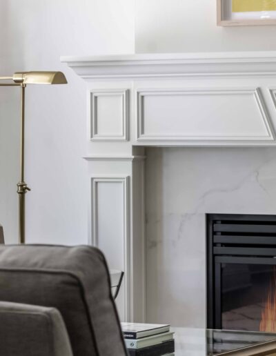 A cozy living room with a lit fireplace, gray armchair, decorative sculpture, standing lamp, and a patterned pillow.