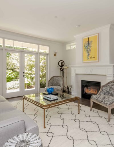 A bright living room with a gray couch, glass coffee table, patterned chairs, and a fireplace. Large windows and a mirror reflect natural light.