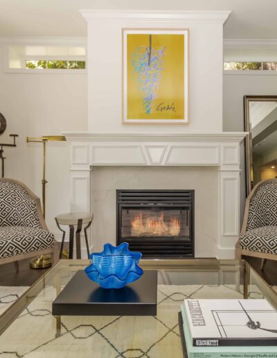 Living room with a fireplace, two patterned chairs, a glass coffee table with a blue decorative bowl, and a framed artwork above the mantel.