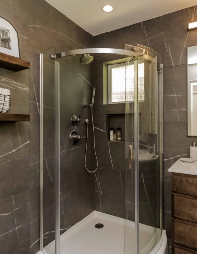 Modern bathroom with a glass-enclosed corner shower, dark tile walls, a white toilet, brown vanity with sink, and shelves holding towels and a plant. Ceiling light and rectangular mirror.