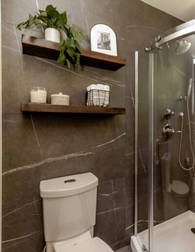 A modern bathroom with dark tiled walls, a glass shower, a toilet, and two wooden shelves holding a plant, candles, and a basket.