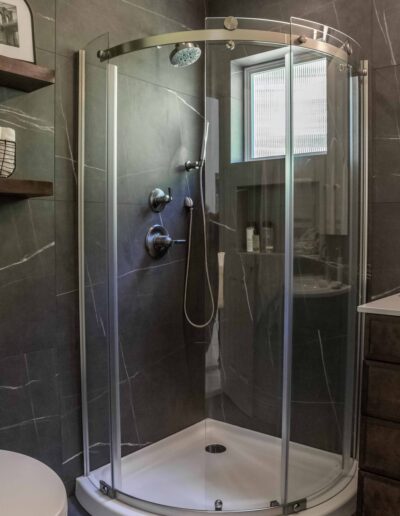 A modern bathroom with a curved glass shower enclosure, dark tiles, and metal fixtures.