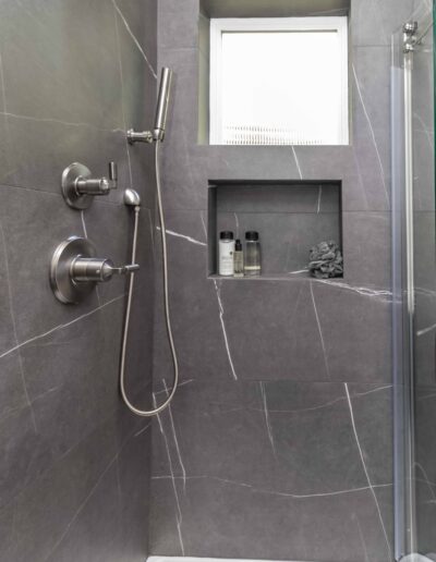 A modern shower with gray marble walls, a rain showerhead, and a handheld shower. Built-in shelf holds shampoo and conditioner bottles.