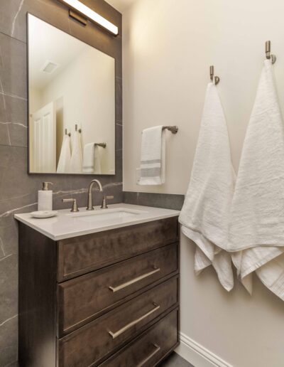 Modern bathroom with a dark wood vanity, rectangular mirror, overhead light, and silver faucet. White towels hang on wall hooks beside the sink.