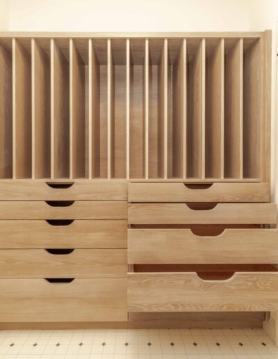 Wooden storage unit with multiple vertical slots above and several drawers below, some of which are partially open, on a tiled floor.