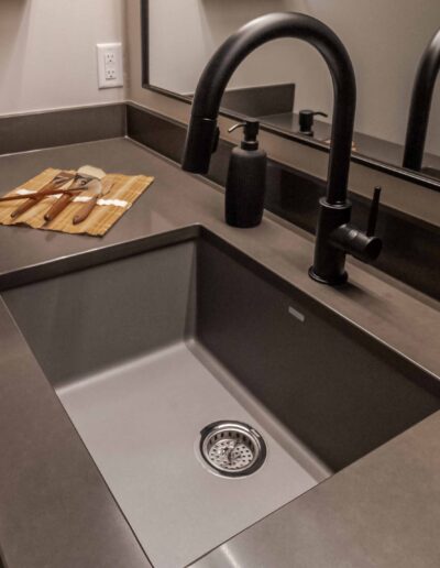 Modern bathroom sink with a black faucet, matching soap dispenser, and a bamboo mat with brushes on the counter. A large mirror reflects the countertop and faucet.