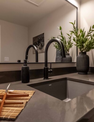 Modern bathroom sink with a black faucet on a dark countertop. A black vase with greenery, a folded gray towel, and a bamboo mat with utensils are neatly arranged on the counter.