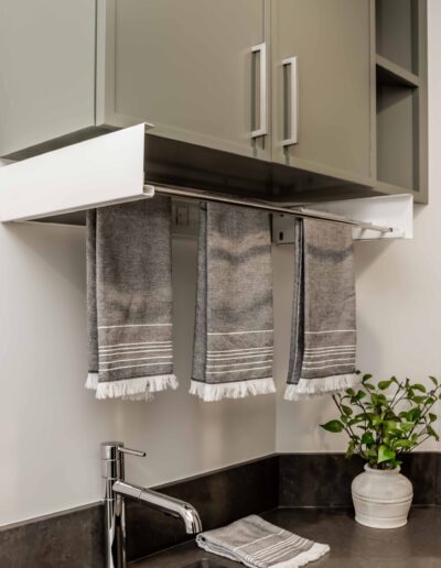 Modern kitchen sink with a silver faucet, gray countertop, and green cabinets. Three gray towels with white fringe hang underneath the cabinet, next to a small potted plant.