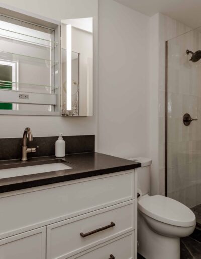 Modern bathroom with a white vanity, black countertop, and single sink. Glass shower enclosure on the right. Decor includes a vase with flowers and a soap dispenser.