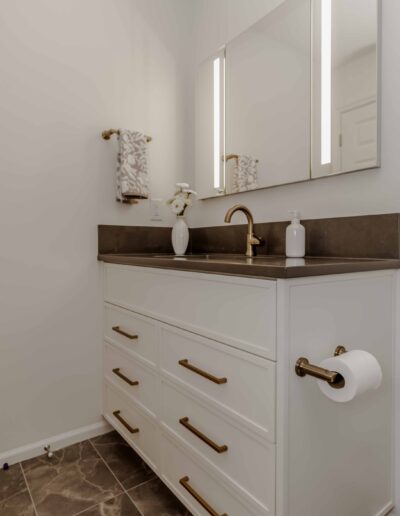 Modern bathroom with a white vanity, brown countertop, and brass fixtures. Features a large mirror, a towel, and a toilet paper holder on the side.