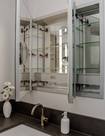 A bathroom vanity with a mirror cabinet open to reveal glass shelves. A white sink and faucet are below, with a soap dispenser and a vase of flowers on the countertop.