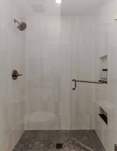 Modern shower with white tiled walls, a round showerhead, a grab bar, and a niche holding shower products. The floor has dark hexagonal tiles and a central drain.