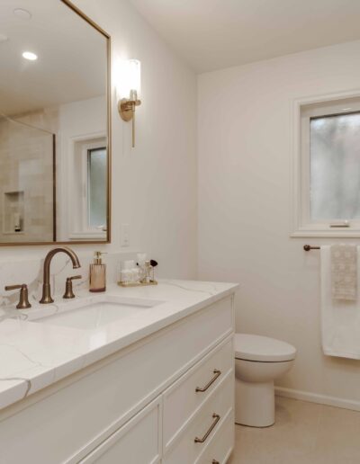 A modern bathroom with a marble countertop, sink, and brass fixtures. There is a mirror, a window, a toilet, and a glass-enclosed shower. Towels are neatly hung on the wall.