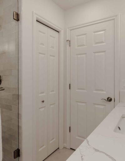 A white bathroom with a marble countertop, a sink, and a towel on a rail. There is a shower to the left and two closed doors with silver handles.