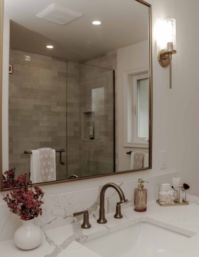 Modern bathroom with a marble countertop, brass faucet, and a large mirror. A vase with flowers and toiletries are placed on the counter. A glass-enclosed shower is visible in the reflection.