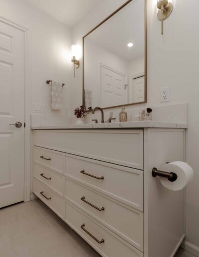 A modern bathroom features a large mirror, a white vanity with brass handles, a countertop with toiletries, wall-mounted lights, and a roll of toilet paper on the side.