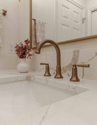 A bathroom sink with a marble countertop, bronze faucet, a beige hand towel on a rack, a vase with flowers, and a pink soap dispenser.