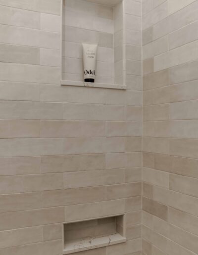 Shower with beige tile walls featuring a shelf holding a tube of Odele shampoo.